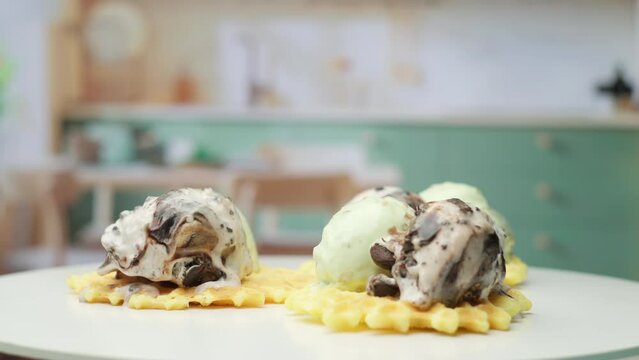 Melting pistachio and chocolate ice ream balls on the top of waffles rotating in the kitchen in slow motion.