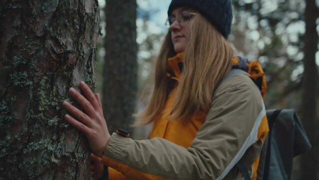 Mountain trekker woman in yellow jacket stops at tree in forest and looks up. Feels the tree's energy while keeping her hand on trunk