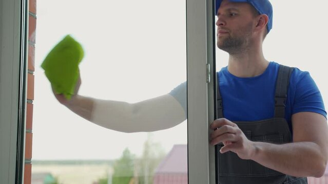 Man worker in blue uniform wipes window glass with green rag from outside. General cleaning in private house. Washing new windows after installation