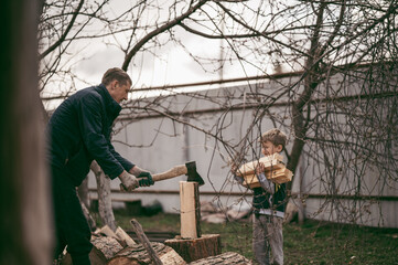 dad is chopping firewood in the yard of a village house, the boy helps dad carry firewood in the woodpile. Dad and son work together