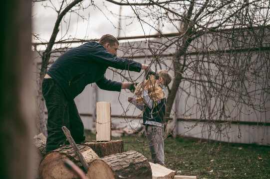 Dad Is Chopping Firewood In The Yard Of A Village House, The Boy Helps Dad Carry Firewood In The Woodpile. Dad And Son Work Together