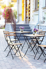 Empty cafe with terrace with tables and chairs. Street exterior of restaurant. Flowers in vase on table. Furniture for coffee shop in street in Europe in summer. Old Cozy street with tables of cafe	