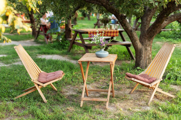 Wooden chairs and table table in summer orchard. Wooden outdoor furniture set for Picnic in garden. Cozy Interior Courtyard with table and sun loungers. Garden furniture for leisure time in nature. 