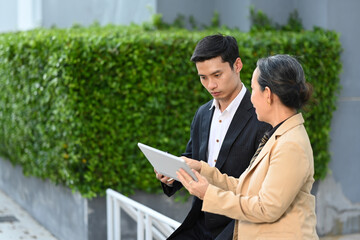 Middle aged woman and young man business people in formalwear discussing online information while sitting outside an office building