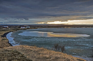 View across lake Myvatn in Iceland, partly frozen under dark clouds with a strip of clear sky and sunlight at the horizon
