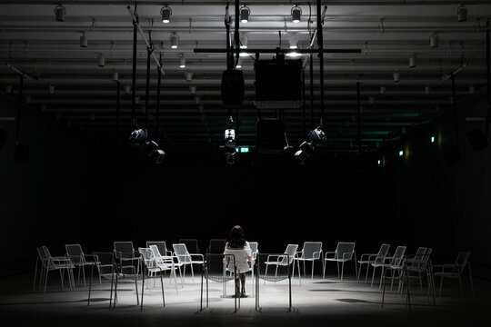 A girl sits in a circle of chairs under a beam of light in a dark room