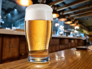 Cooled glass of beer on the wooden table. Blurred bar interior at the background.