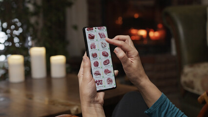 Ordering food using a smartphone at home. A woman selects meat and fish in an online store using an application on a smartphone. Home evening furnishings with a burning fire in the fireplace.