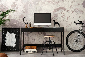 Concrete interior of home office with black desk, computer screen, office accessories, lamp. Rack with personal accessories. Home decor. Template.