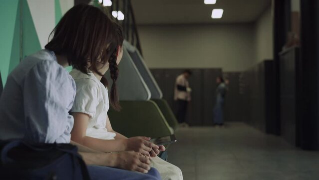 Two schoolgirls smile holding smart pad in school hallway. Kids enjoying break.
