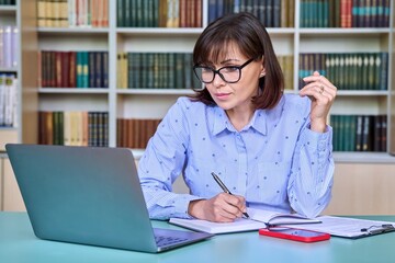 Middle-aged female teacher working in library, using laptop