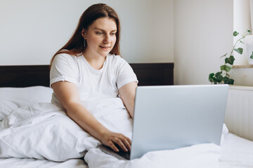 Portrait of beautiful young redhead woman with laptop on bed on a lazy day. Online education, working or shopping. Technology woman concept for alternative office freelance