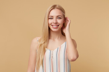 Studio portrait of young blonde woman posing over beige background touching her hair and smiling