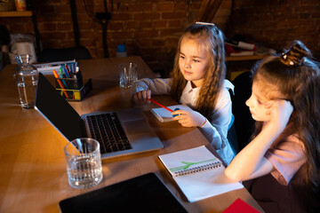Two little cute kids, sisters sitting at home and doing homework at home interior at evening time. Online education, childhood, family, remote learning and school concept.