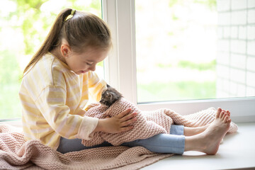 A cute girl plays with her little fluffy kitten sitting on the windowsill of the house. Friendship...