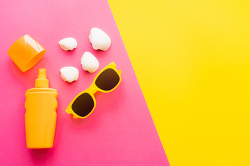 Top view of sunblock and sunglasses near seashells on pink and yellow background.