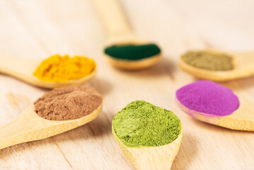 Superfood powders: sweet potato, matcha, spirulina, hemp, turmeric, carob in wooden spoons on a board.
