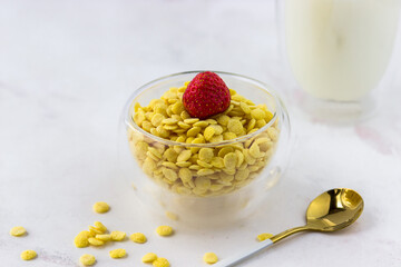 A bowl of breakfast cereal and a glass of milk on a white marble table. Instant corn flakes with strawberries