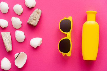 Top view of seashells near sunglasses and bottle of sunscreen on pink background.