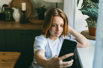 A teenage girl with red hair sits near the window and looks into the phone. 
