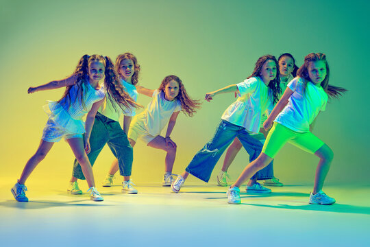 Group of children, little girls in sportive casual style clothes dancing in choreography class isolated on green background in yellow neon light. Concept of music, fashion, art