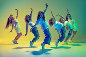 Groep kinderen, kleine meisjes in sportieve casual stijl kleding dansen in choreografie klasse geïsoleerd op groene achtergrond in geel neonlicht. Concept van muziek, mode, kunst