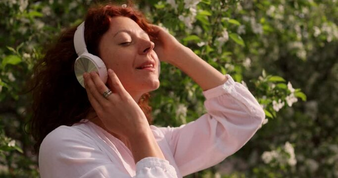 Red-haired woman in headphones in a flowering garden listening to music while dancing