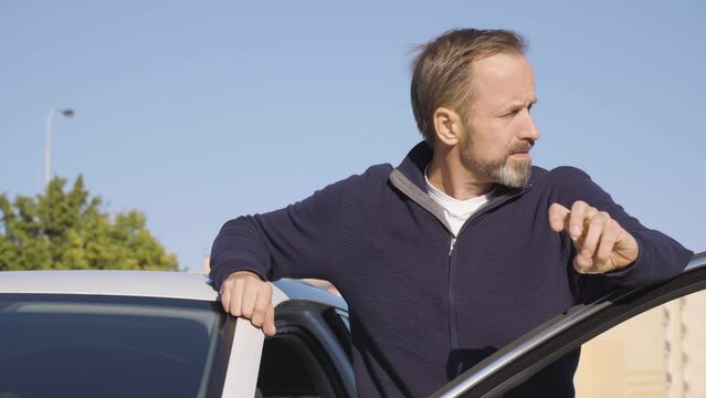A middle-aged handsome Caucasian man looks around in thought as he stands next to his car