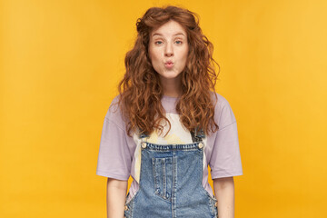 indoor portrait of young ginger curly female posing over yellow bright background showing silence gesture and looks aside at copy space