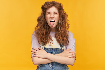 indoor portrait of young ginger curly female posing over yellow bright background showing her tongue into camera