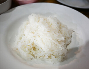 Thai vermicelli (Khanom Chin or slightly fermented noodles) served on a white plate with a spoon and fork. The most popular menu is Kanom Jeen, Fish Curry with Vegetables or Spicy Khanom Jeen Salad.