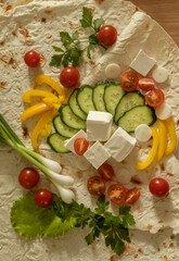Still life with cherry tomatoes, cucumbers, peppers, herbs, onions and cheese spread on a tortilla on a wooden table top view