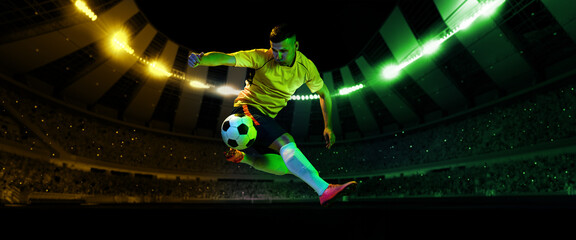 Energetic professional soccer player jumping with football ball at crowded stadium with flashlights at evening time. Concept of sport, competition, motion, overcoming.