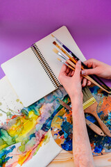 Flat lay view on open blank sketchbook, watercolor drawing, brushes, palette. The girl holds brushes in her hands.