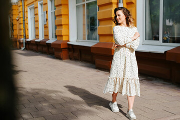 Charming young woman with curly hair and white polka dot dress walking down street in city. Pretty...
