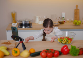 Young Girl Vlogger Making Social Media Video About Cooking At Home, looking on smartphone on tripod. episode about healthy eating. concept of attraction children to healthy diet of early age