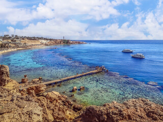Fototapeta premium View from the cliff to the beach with a coral reef in Sharm El Sheikh, Egypt. Wooden pier among the stones in the Red Sea and two ships on the horizon