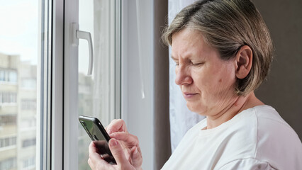 Old woman scrolls through web pages on black smartphone standing near window. Woman in white t-shirt checks social media on mobile phone