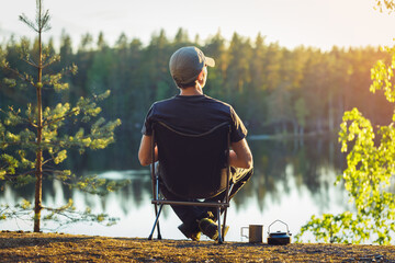 A man is sitting in a camping chair on a summer evening on the background of a forest lake. - obrazy, fototapety, plakaty
