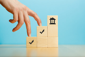 Fingers of hand go up the steps of the pyramid of wooden blocks with house and marks icons on blue background.