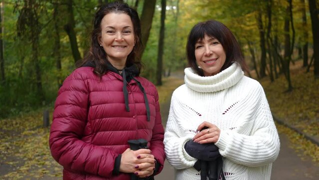 Medium shot portrait of two happy mature women looking at camera smiling standing on alley in autumn park. Confident attractive Caucasian friends posing outdoors with ski poles. Sport and friendship
