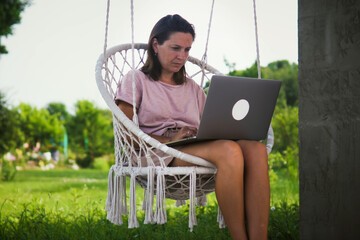 Young woman using laptop computer while sitting in cozy garden swing chair at backyard of her...