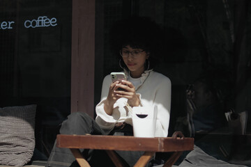 Beautiful smiling african american woman texting with friends using her mobile phone and eats a street food at summer day. Woman with afro hair eats her lunch outdoors and using her mobile phone