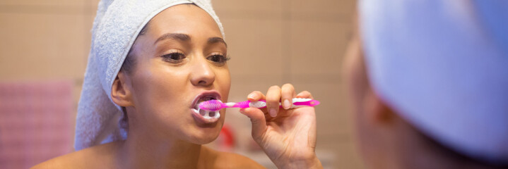 Smiling young woman brushing teeth with toothbrush in front of the bathroom mirror