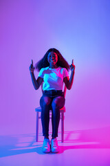 Fototapeta na wymiar Cool promo. Young black lady sitting on chair, pointing upwards, offering empty space in neon light, mockup for ad