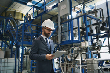 Chief Engineer in the hard hat walks through industrial factory while holding tablet.