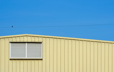 Aluminum louver on yellow corrugated steel warehouse wall with little bird perching on electric line against blue sky background