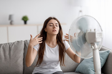 Sad young european woman suffering from heat sits on sofa, catches cold air from fan and waves hands to herself
