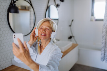 Beautiful senior woman in bathrobe listening to music in bathroom, relax and wellness concept. - obrazy, fototapety, plakaty
