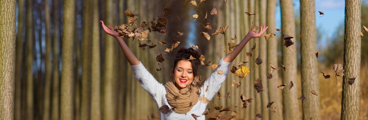Beautiful smiling girl throws falling dry forest leaves in the air on a sunny autumn day
